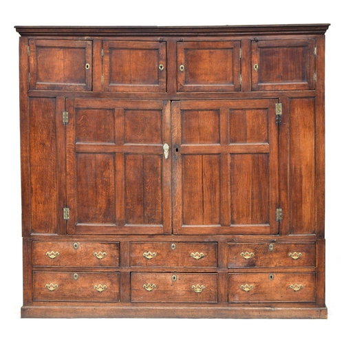 A massive 18th century oak housekeeper's cupboard, in two sections, the top with four cupboards, over a pair of panelled doors opening to two well worn pine shelves, the interior with a painted boarded back, the base with an arrangements six cockbeaded drawers, on a plinth, 214cm wide, 51.5cm deep, 202cm high