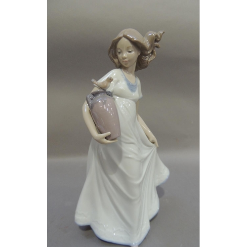52 - A Nao porcelain figure of a young girl carrying a two handled vase under her arm with a bird perched... 
