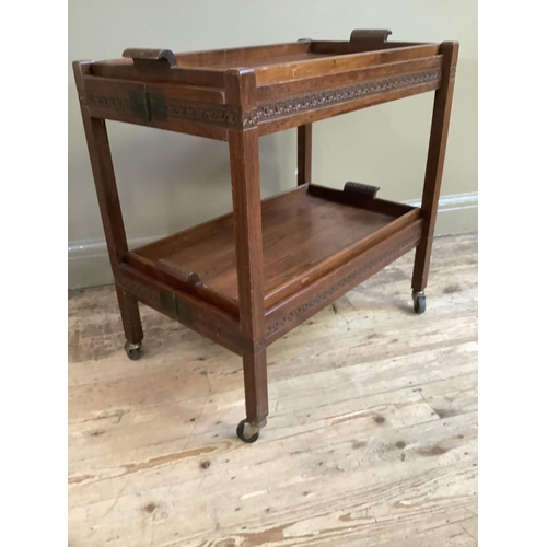 323 - A folding wooden tea trolley of two tiers with removable trays carved with a band of stylised flower... 