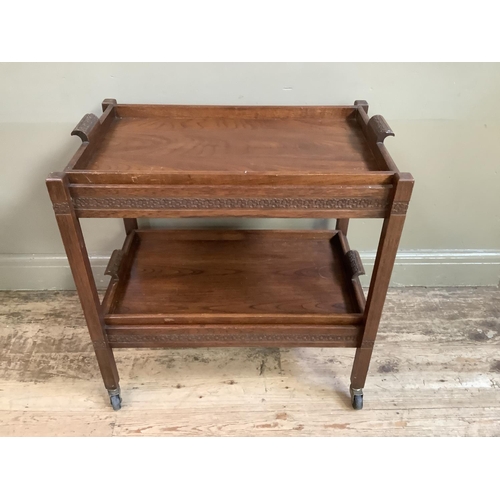 323 - A folding wooden tea trolley of two tiers with removable trays carved with a band of stylised flower... 