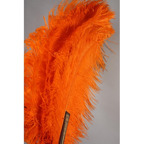 101 - An orange ostrich feather fan, the monture of patterned wood, the guards decorated in gold, overall ... 