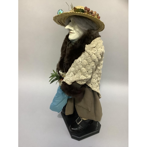 23 - A figure of an elderly woman wearing a straw hat, faux fur coat with shawl carrying umbrella and var... 