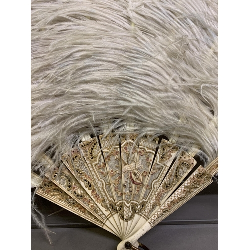 109 - A large white ostrich feather fan, the bone sticks carved, pierced and coloured in several shades of... 