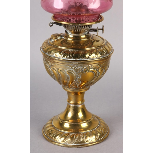 15 - A VICTORIAN BRASS OIL LAMP, with rocaille and scroll reservoir, on conforming base, cranberry glass ... 