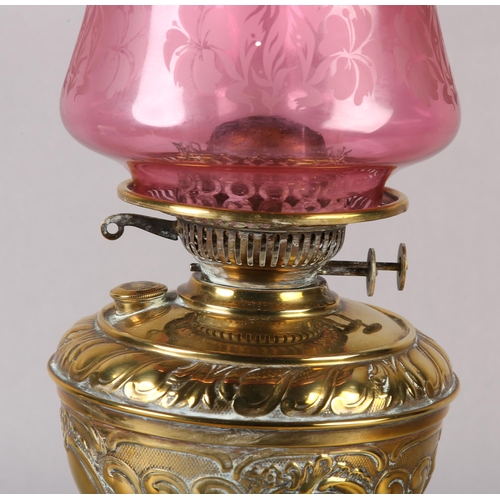 15 - A VICTORIAN BRASS OIL LAMP, with rocaille and scroll reservoir, on conforming base, cranberry glass ... 