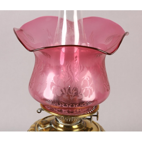 15 - A VICTORIAN BRASS OIL LAMP, with rocaille and scroll reservoir, on conforming base, cranberry glass ... 