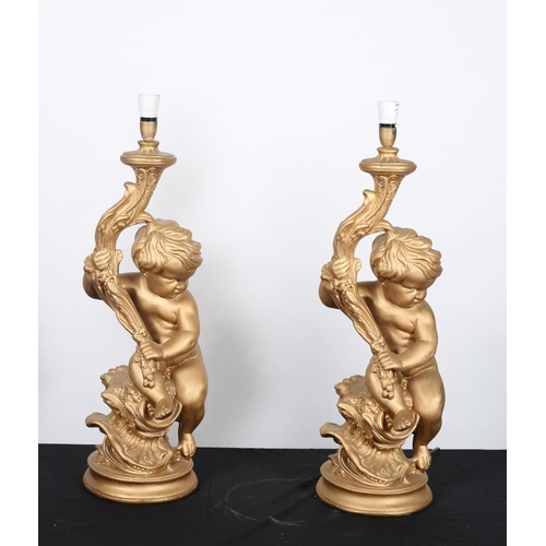 563 - A PAIR OF VINTAGE GILT PLASTER FIGURAL TABLE LAMPS each modelled as putti shown seated holding a tor... 