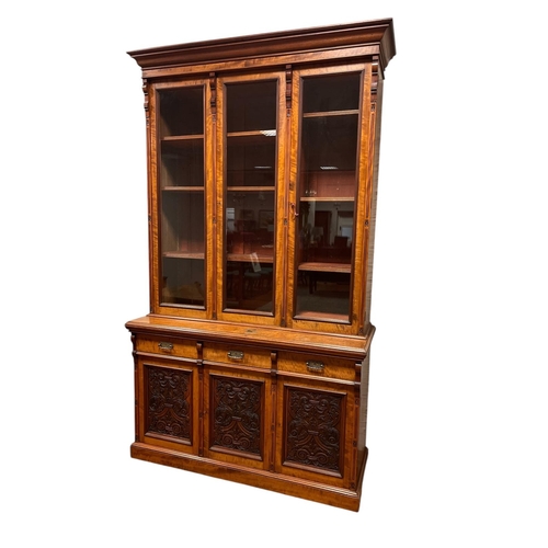 AN EDWARDIAN SATIN BIRCH LIBRARY BOOKCASE the moulded cornice above three glazed doors containing adjustable shelves the base with three frieze drawers and brass drop handles above three cupboards with carved panels on platform support 266cm (h) x 155cm (w) x 60cm (d)