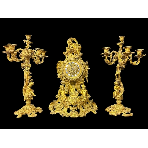 A FRENCH 19TH CENTURY GILT BRONZE THREE PIECE CLOCK GARNITURE comprising a large Rococo style mantel clock of waisted form surmounted by a reclined putto the dial set with blue and white enamel Roman numerals above further opposed figural putti and intricately rendered rocaille motifs throughout the pierced splayed flower strewn base on scrolled feet together with two six light candelabra with flowering sconces on scrolled vegetal arms and conforming stems putti and bases Clock 62cm (h) x 46cm (w) x 24cm (d) (3) (AF)