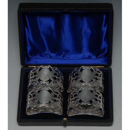 593 - A set of four Edwardian silver demi-lune napkin rings, each pierced with scrolls and centred by a va... 