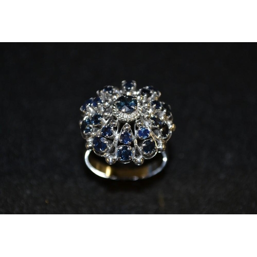 3436 - A sapphire cluster ring, round floral shield crest inset with twenty one blue sapphires, white gold ... 