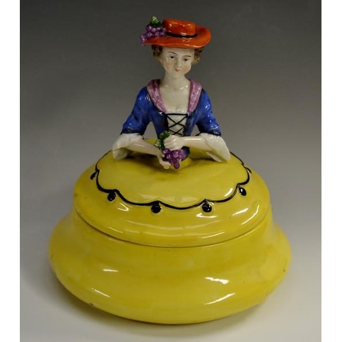 3114 - A Sitzendorf powder bowl and cover, modelled as a lady in 18th century dress holding a bunch of grap... 