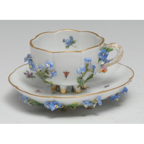 760 - A Meissen lobed cabinet cup and saucer, painted with flowers, the exteriors encrusted, six spur feet... 