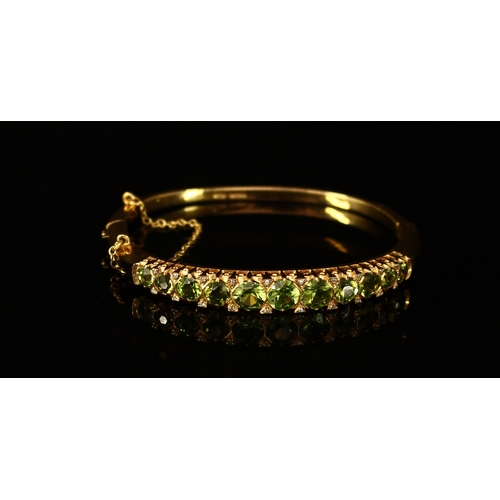 303 - A 9ct gold and peridot hinged bangle, the 11 graduated stones divided by pairs of small brilliant cu... 