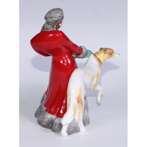 634 - A Royal Crown Derby figure, Olga, she stands, wearing a fur trimmed red trench coat holding a Borzoi... 