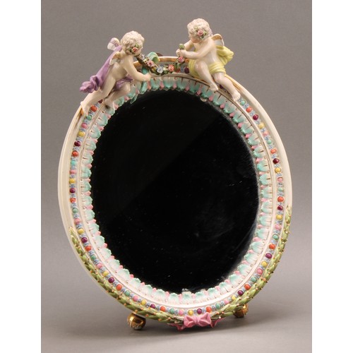 229 - A German porcelain looking-glass, oval mirror plate, the frame crested by a pair of cherubs, the bor... 