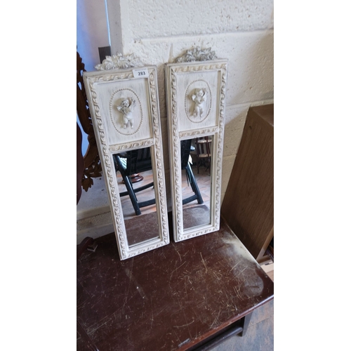 283 - Pair of vintage white-framed mirrors with cherub relief designs, intricate floral carvings, and beve... 