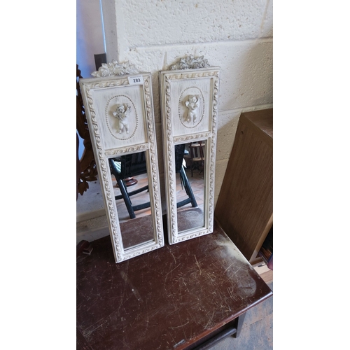 283 - Pair of vintage white-framed mirrors with cherub relief designs, intricate floral carvings, and beve... 