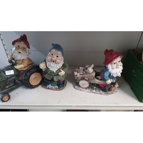 129 - Set of three ceramic garden gnome figurines: one on a tractor, one with a cane, and one pushing a wh... 