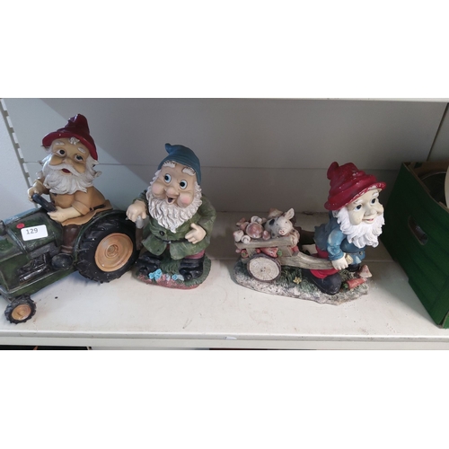 129 - Set of three ceramic garden gnome figurines: one on a tractor, one with a cane, and one pushing a wh... 
