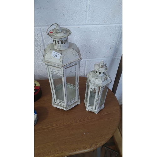 339 - Pair of distressed white metal lanterns with glass panels. One larger lantern paired with a smaller ... 