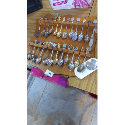 36 - Collection of 24 souvenir spoons with diverse enamel crests on a wooden rack, featuring various moti... 