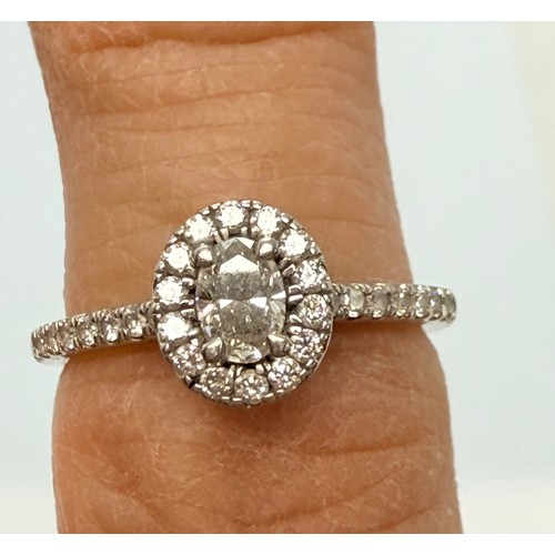  A platinum diamond engagement ring, the central oval brilliant cut stone enclosed by a halo of round... 