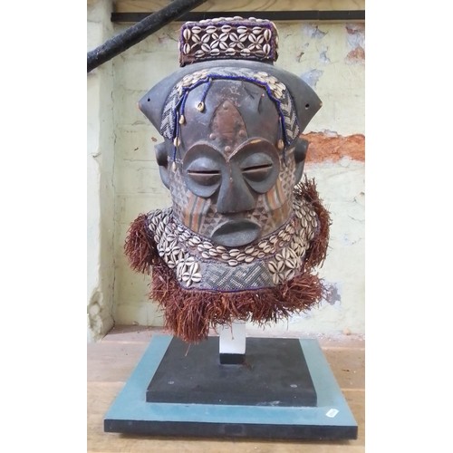 138 - An African carved wood mask painted and decorated with shells, on stand, height 61cm.