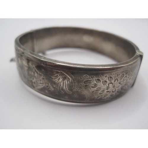 13 - A hinged silver bangle with box snap catch and safety chain,
band width 16mm, half-engraved with flo... 