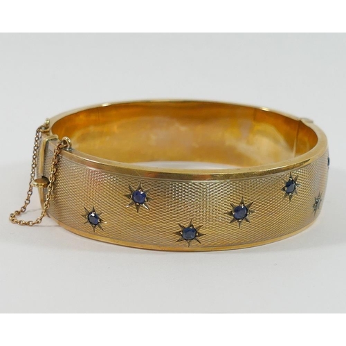 178 - A 9 carat gold hollow hinged sapphire set bangle, with engine turned decoration, the seven round mix... 