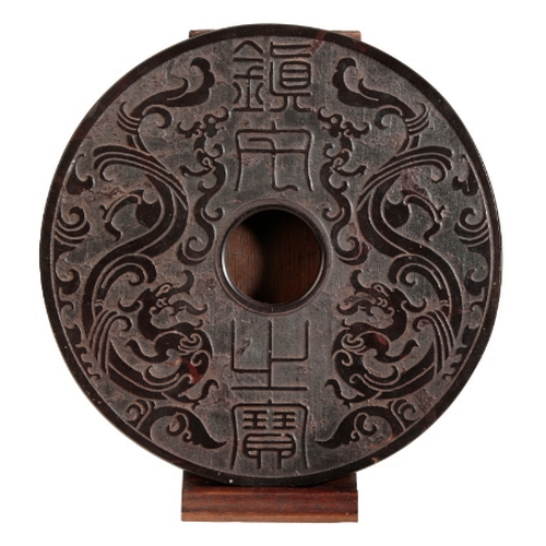 56 - A LARGE CHINESE BLACK HARDSTONE BI carved with archaistic dragons, 30cm diameter, on a wooden stand