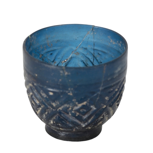 219 - AN ISLAMIC BLUE GLASS VESSEL with moulded geometric decoration and a low circular foot, 9.75cm high ... 