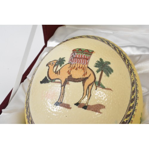 119 - Cased ostrich egg, hand applied decoration depicting camel / animals hunting scenes