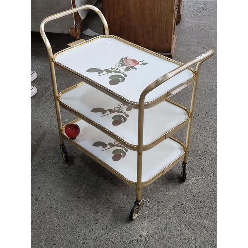 874 - A charming vintage drinks trolley with gold toned frame and three tiers boasting rose ornament. The ... 