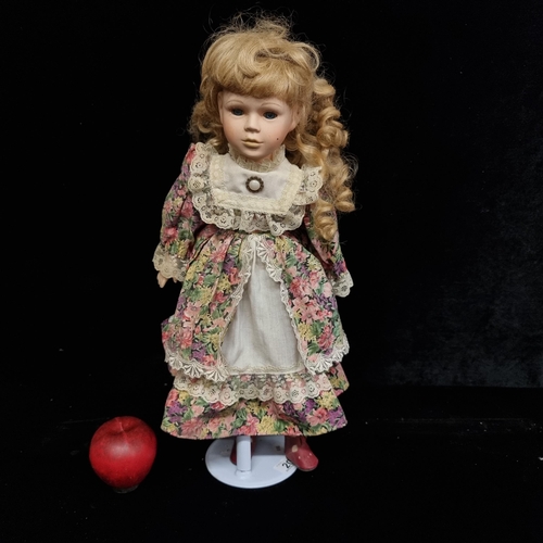 A traditional porcelain doll, dressed in a colorful Edwardian style dress with blonde ringlets and accompanied by a stand.