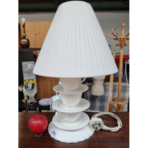 468 - A whimsical table lamp with a figural teacup and saucer base in white porcelain. Topped with a pleat... 