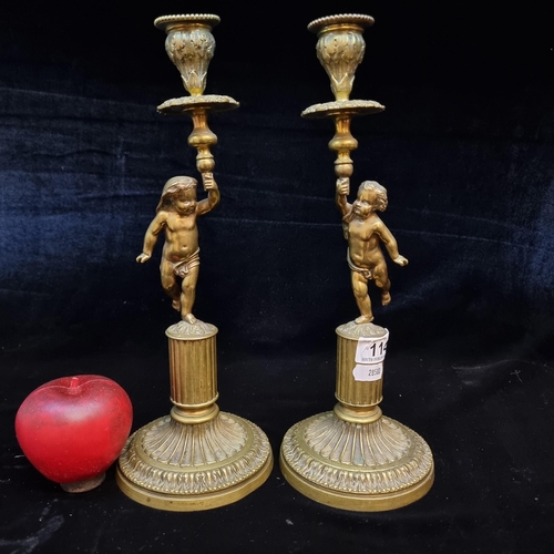 114 - A pair of antique heavy brass candlesticks surmounted with figural cherub / putti tops. Supported by... 