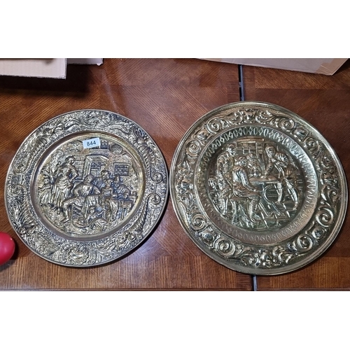 844 - Pair of large decorative brass wall plates featuring embossed scenes of village life, adorned with i... 