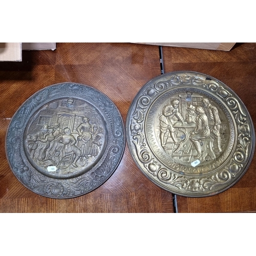 844 - Pair of large decorative brass wall plates featuring embossed scenes of village life, adorned with i... 