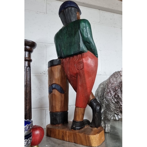 986 - A large heavy hand-carved wooden statue of a man leaning against a wooden plinth. Painted in vibrant... 