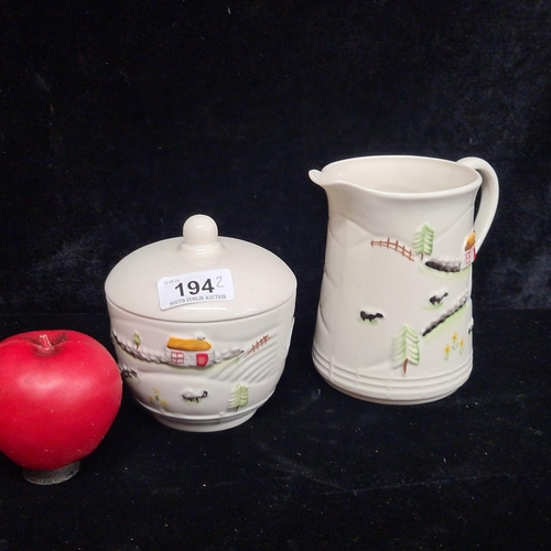 194 - A Belleek Irish milk jug and lidded sugar jar. Both featuring a wholesome rural scene of a cottage a... 