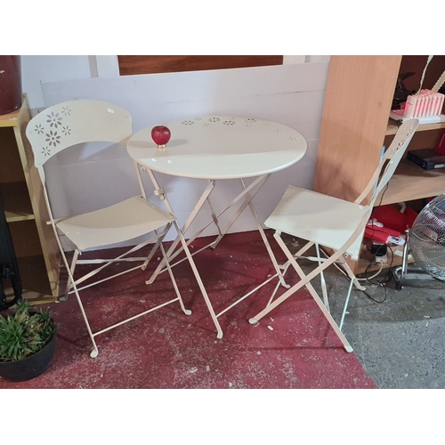 923 - Cream metal folding bistro set includes a round table and two matching chairs with decorative cut-ou... 