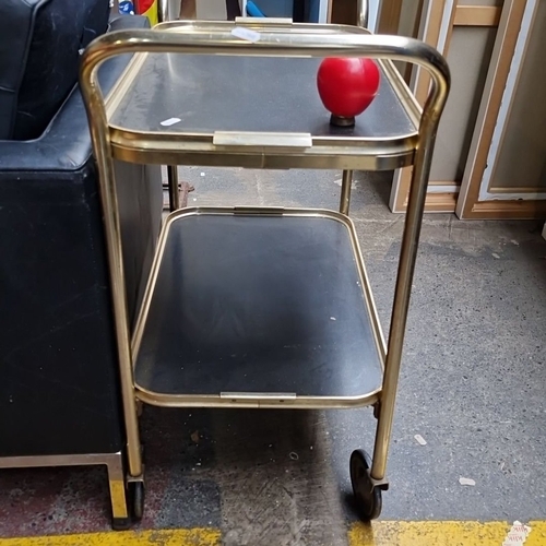 638 - Mid-century modern two-tier bar cart with brass-tone metal frame and black trays. Features smooth-ro... 