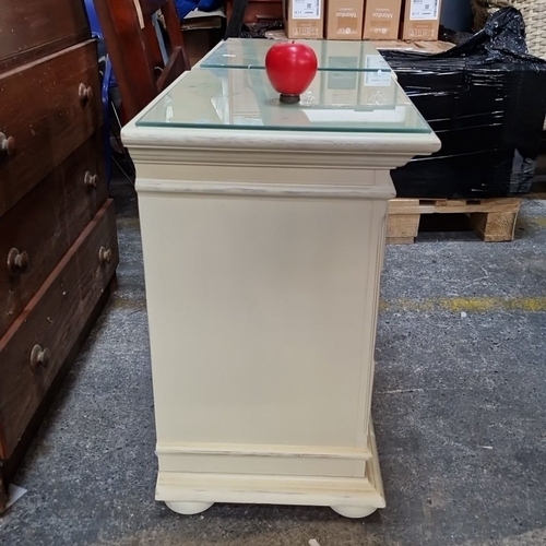 1162 - A Pair of vintage cream-painted wooden bedside cabinets with three drawers each, glass tops, and cla... 