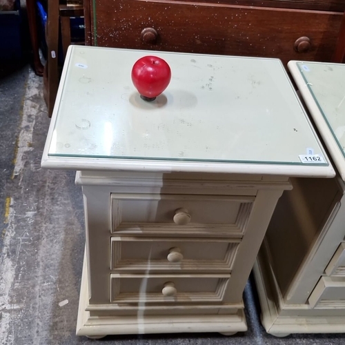 1162 - A Pair of vintage cream-painted wooden bedside cabinets with three drawers each, glass tops, and cla... 