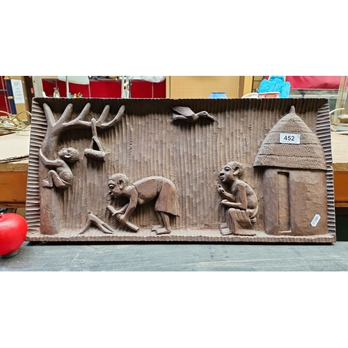 Hand-carved tribal relief panel depicts daily village life, detailed with figures, a hut, and wildlife, crafted from solid wood with textured accents.