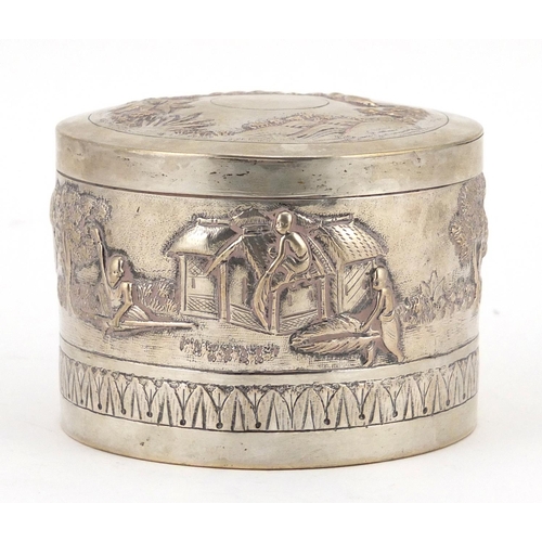 374 - Indian unmarked silver cylindrical box and cover embossed with figures in a village, 7cm high x 9cm ... 