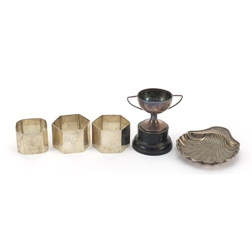 384 - Silver objects comprising three napkin rings, shell shaped dish and miniature trophy with twin handl... 