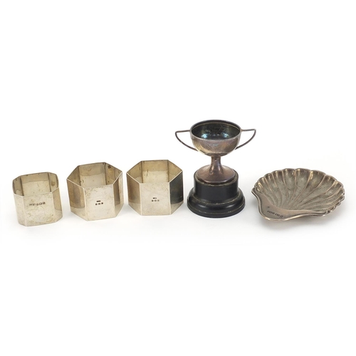 384 - Silver objects comprising three napkin rings, shell shaped dish and miniature trophy with twin handl... 