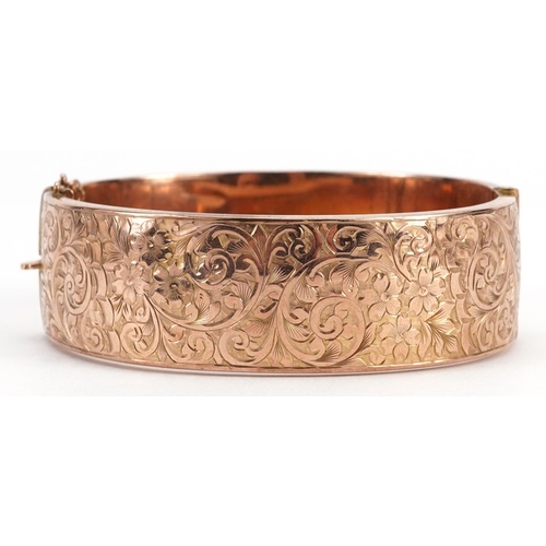 2058 - 9ct rose gold hinged bangle with engraved floral decoration and safety chain, 6.3cm wide, 20.1g